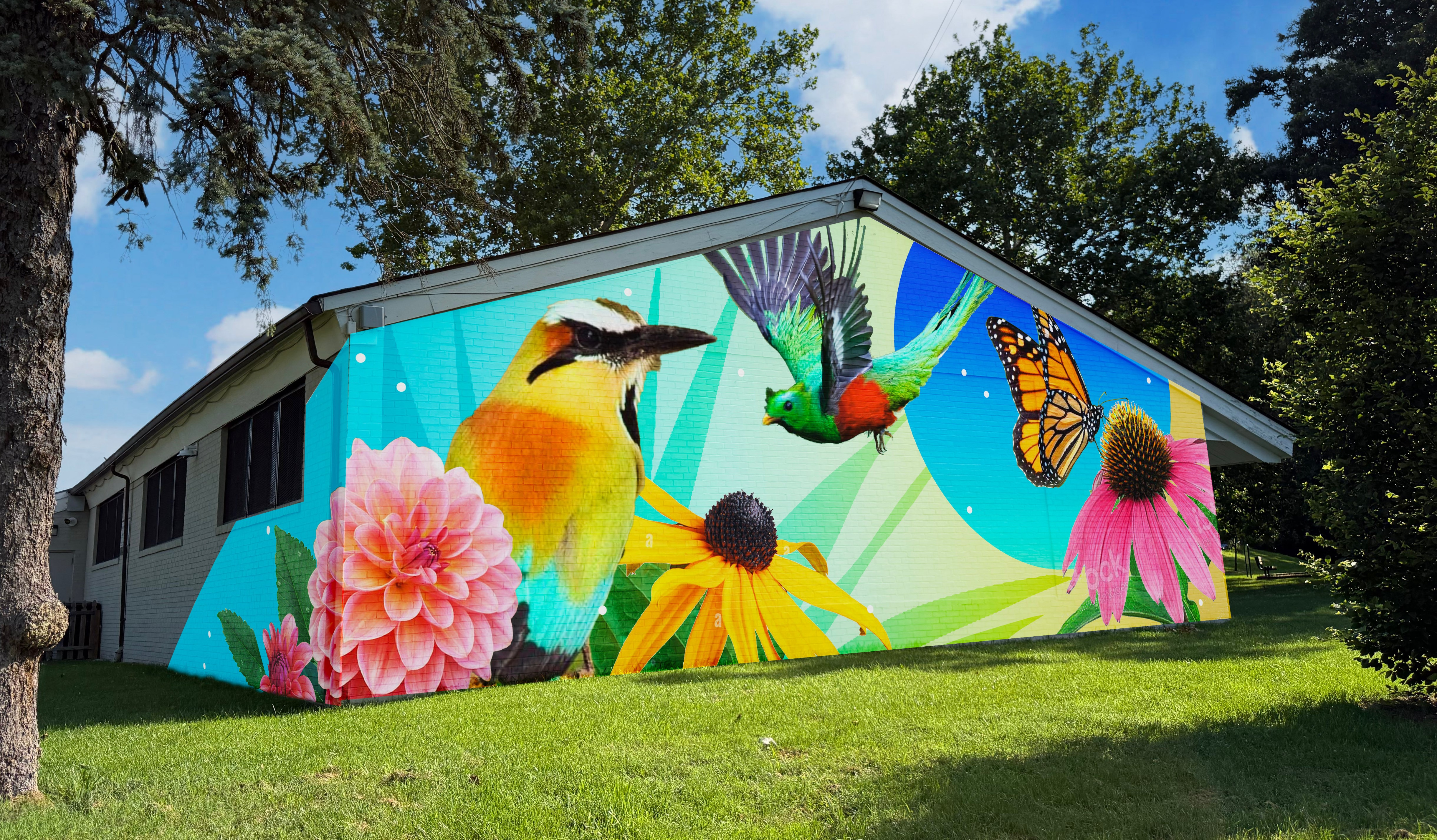 Mural design concept titled Rooted and Radiant. The Image is a rendering of the front-facing wall with birds, a butterfly, and flowers, set to a blue and green background